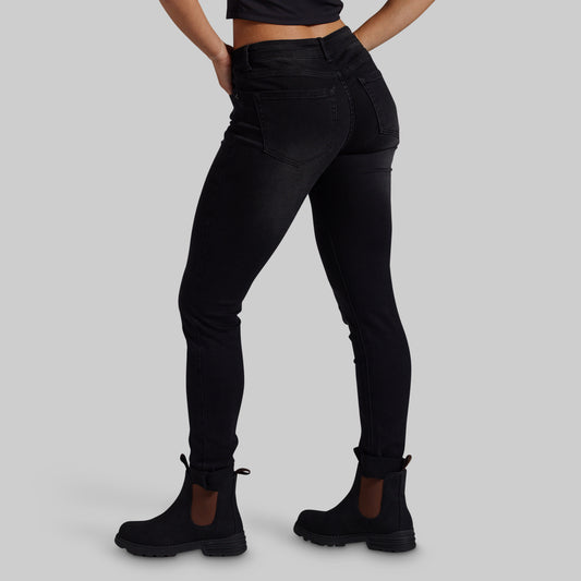 Rear three-quarter view of a model wearing black high-waisted skinny jeans and black Chelsea boots against a solid gray studio background.