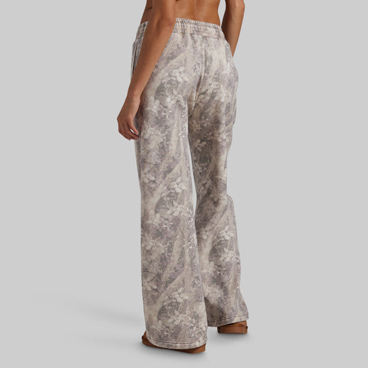 A person shown from the back wearing wide-leg pants with an abstract floral or marble-like print in beige, taupe, and gray tones, against a plain light gray studio background.