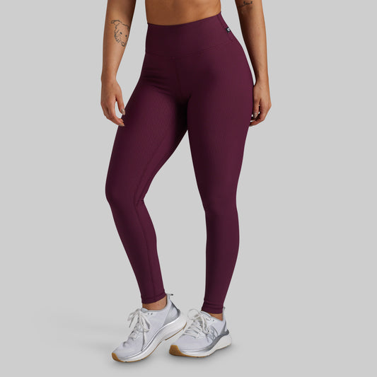 A model wearing high-waisted ribbed leggings in a deep plum purple color, paired with white athletic sneakers, shown from the waist down against a solid light gray background.