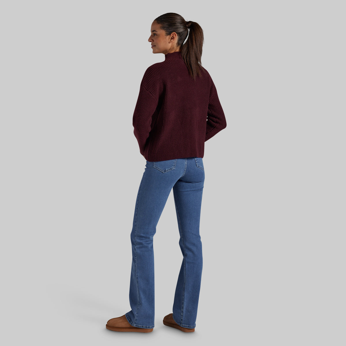 A woman shown from the back wearing a dark burgundy ribbed mock-neck sweater, blue jeans, and brown mule shoes against a solid gray studio background.