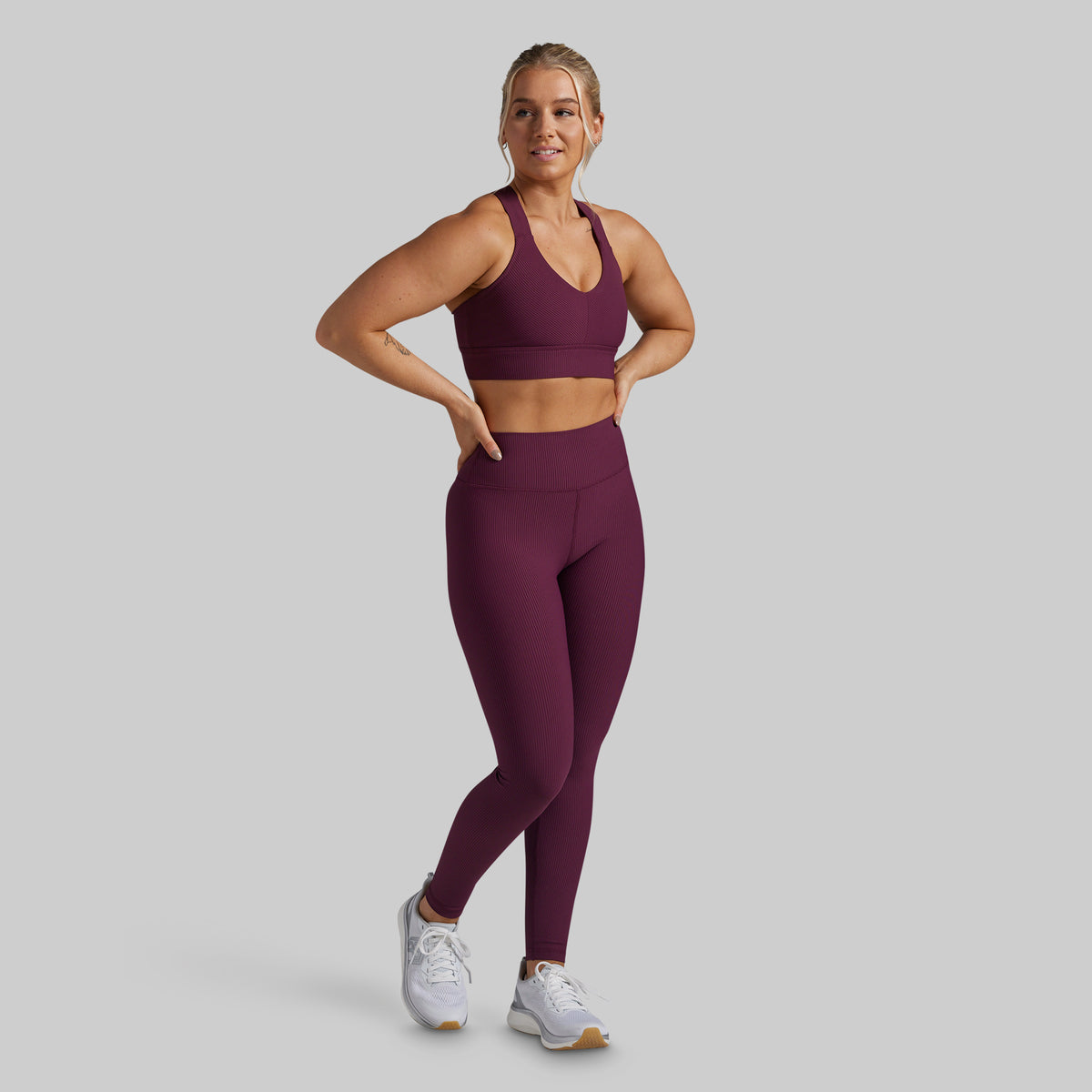 A woman modeling a matching athletic set consisting of a scoop-neck sports bra and high-waisted leggings in a deep plum purple color, paired with white sneakers, against a solid light gray background.