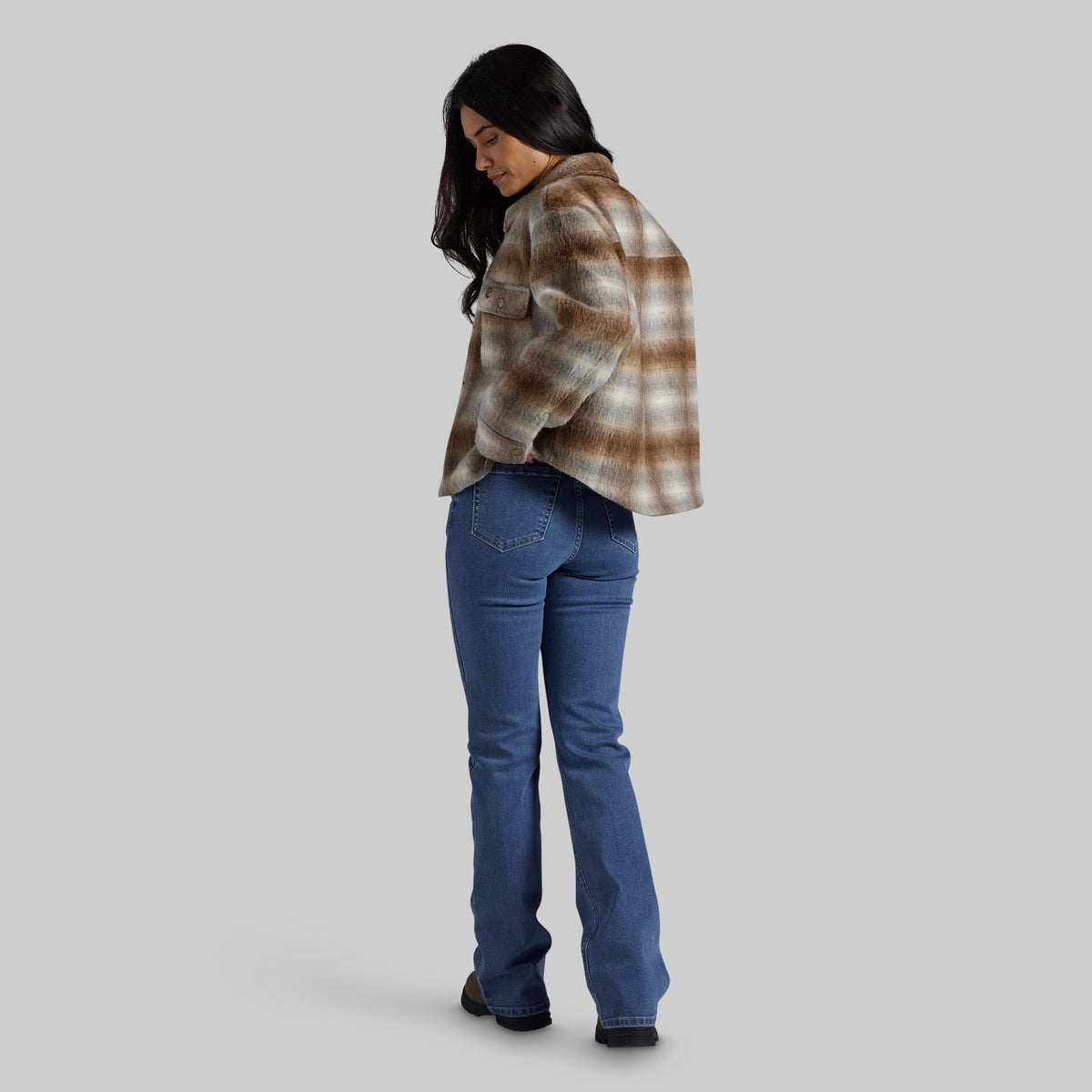 A woman wearing a brown and beige plaid cropped flannel jacket and blue jeans, shown from a side-back angle against a solid gray background.