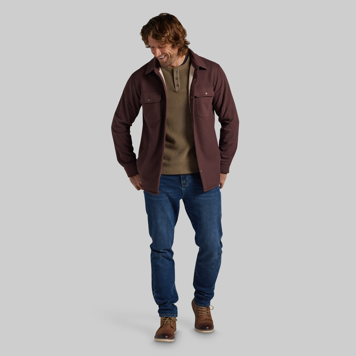 A man modeling a dark brown jacket over a brown henley sweater, paired with blue jeans and brown shoes, against a solid gray background.