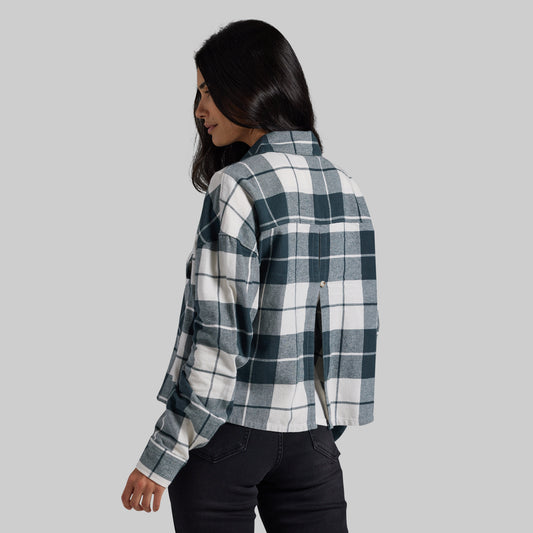 A woman shown from the back wearing a long-sleeve plaid button-up shirt in dark teal and white, paired with black jeans, against a solid light gray background.