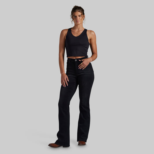 A woman modeling a black sleeveless crop top and black mid-waisted bootcut jeans against a solid light gray background.