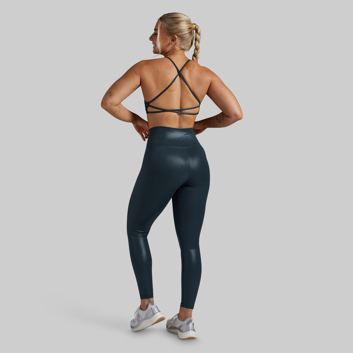 A woman is shown from the back wearing a dark teal athletic sports bra with crisscross straps and matching high-waisted, shiny leggings, paired with gray and white sneakers, against a solid gray background.