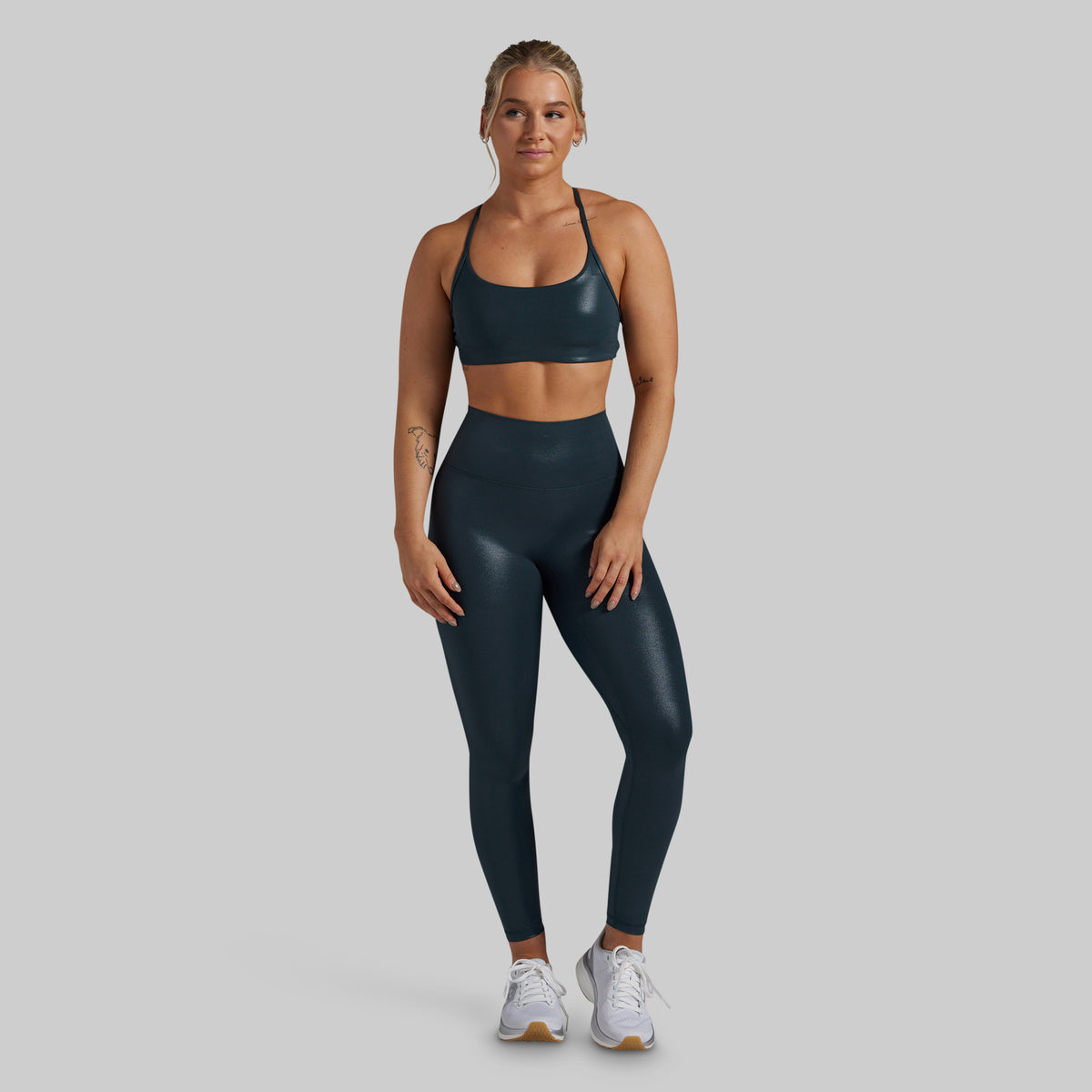 A woman modeling a matching dark teal athletic set consisting of a sports bra and high-waisted leggings, paired with white sneakers, against a solid light gray background.
