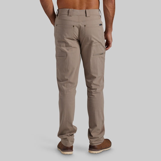 A man viewed from the back wearing a pair of light brown cargo pants and brown shoes, set against a solid gray studio background.