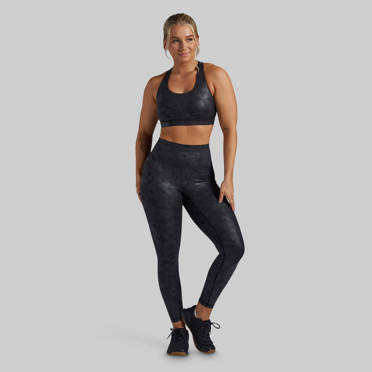 A woman modeling a matching black activewear set, consisting of a sports bra and high-waisted leggings, against a solid gray background.