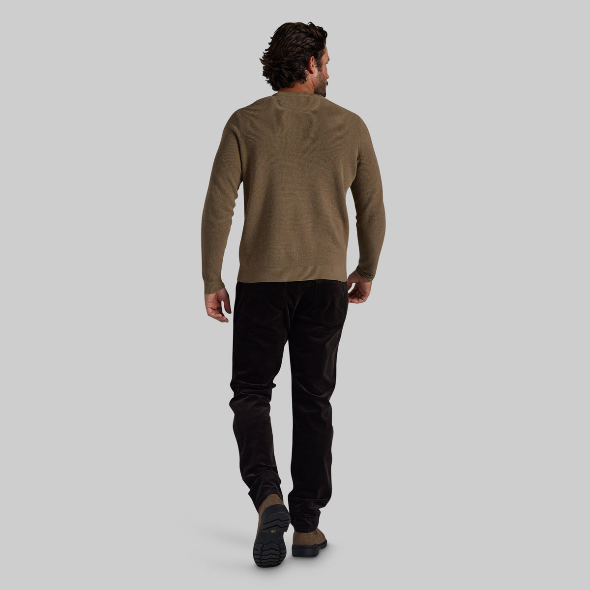 A man viewed from behind wearing a brown knit sweater and black pants, walking against a solid light gray background.