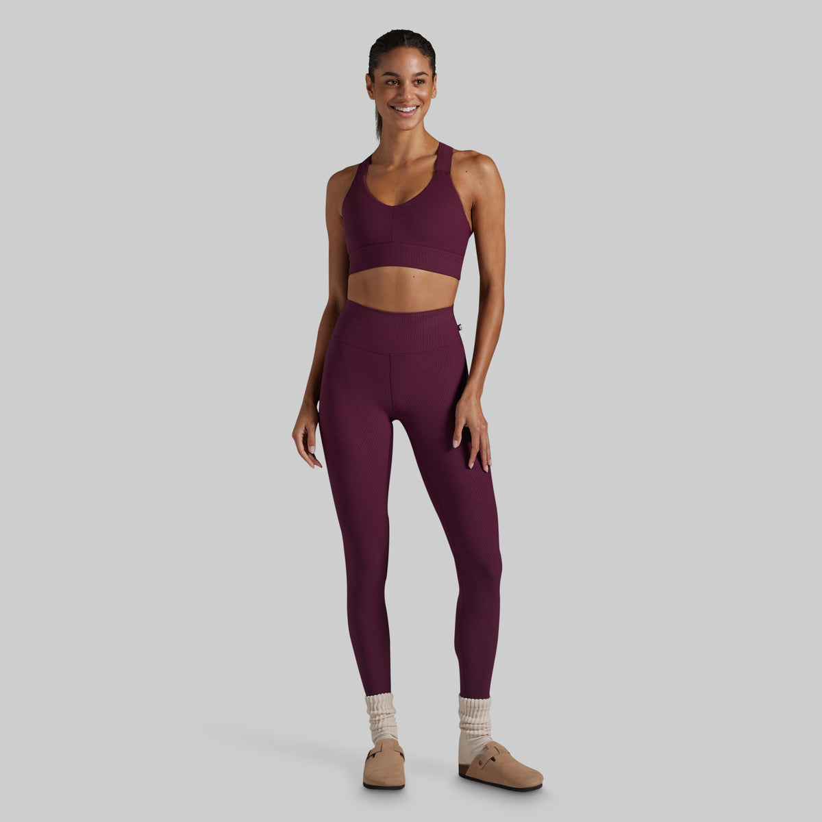 A woman modeling a matching athletic set consisting of a scoop-neck sports bra and high-waisted leggings in a deep plum purple color, styled with beige socks and tan shoes against a solid gray background.