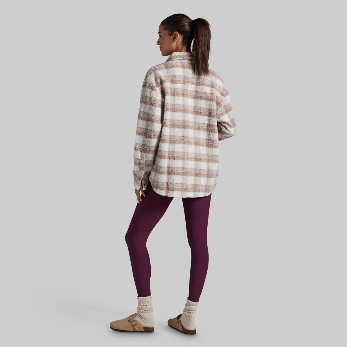 A woman shown from the back wearing an oversized plaid button-up shirt in cream and brown, paired with dark purple leggings, cream socks, and tan slip-on shoes, against a solid gray background.