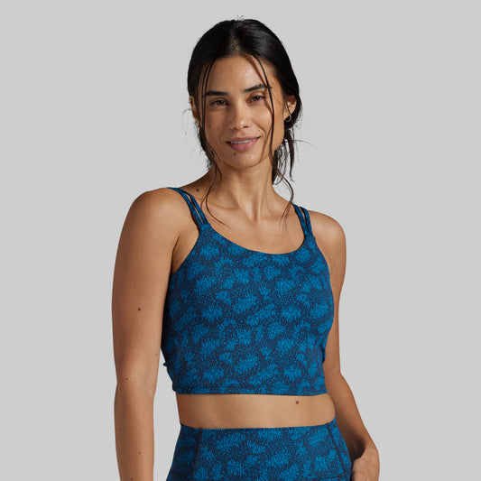 A woman modeling a blue patterned sports bra and matching high-waisted bottoms against a solid gray studio background.