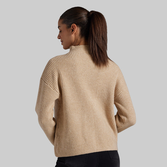 A woman shown from the back wearing a ribbed knit mock neck sweater in a warm beige color, paired with black pants, against a solid light gray background.