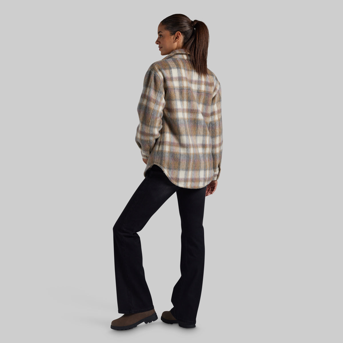 A woman is shown from the back wearing a beige and brown plaid flannel jacket and black pants, standing against a solid gray studio background.