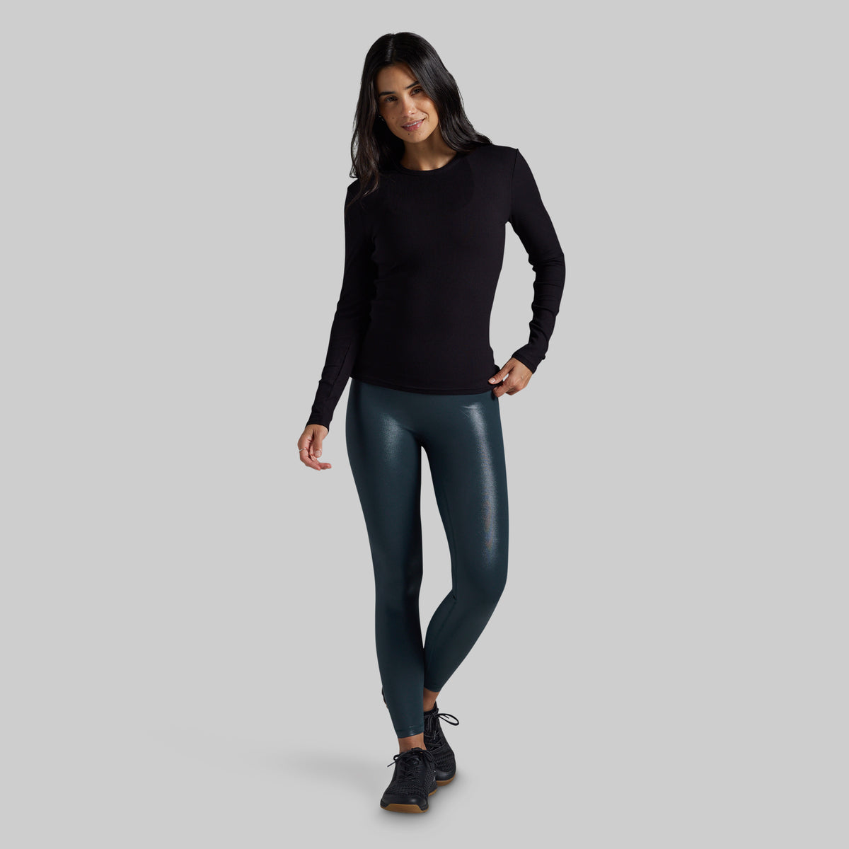 A woman modeling a black long-sleeve crewneck top and dark teal faux-leather leggings with black sneakers, standing against a solid gray studio background.