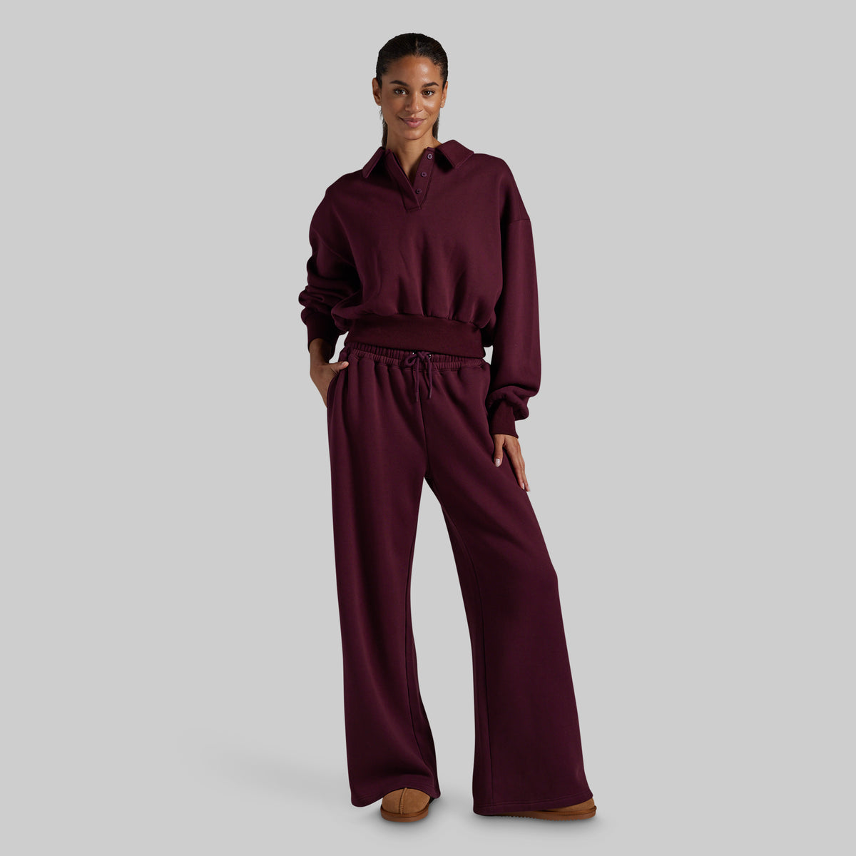 A model wearing a matching burgundy two-piece loungewear set consisting of a cropped long-sleeve collared sweatshirt and wide-leg drawstring sweatpants, set against a solid light gray background.