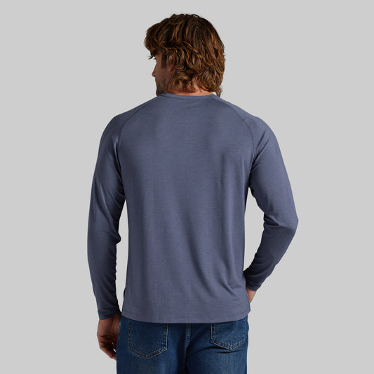 A man viewed from behind wearing a slate blue long-sleeve raglan t-shirt and blue jeans, set against a solid light gray background.