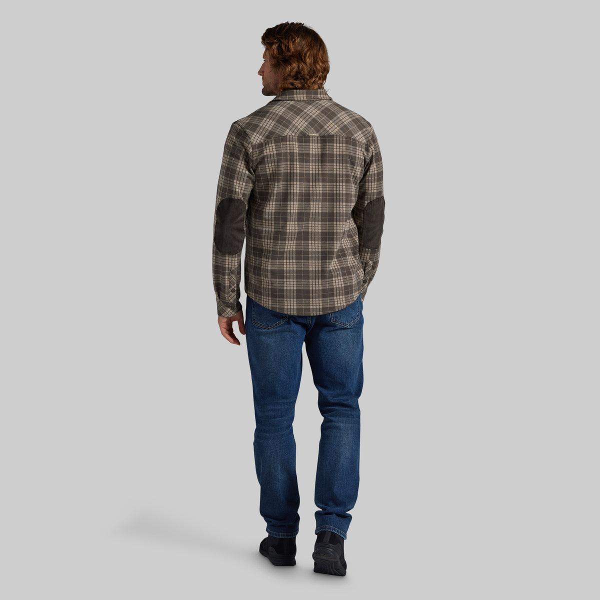 A man viewed from behind wearing a brown and beige plaid button-up shirt with dark brown elbow patches, blue jeans, and black boots, set against a solid gray background.