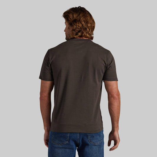A man viewed from the back wearing a dark grey short-sleeve t-shirt and blue jeans, standing against a solid light gray background.
