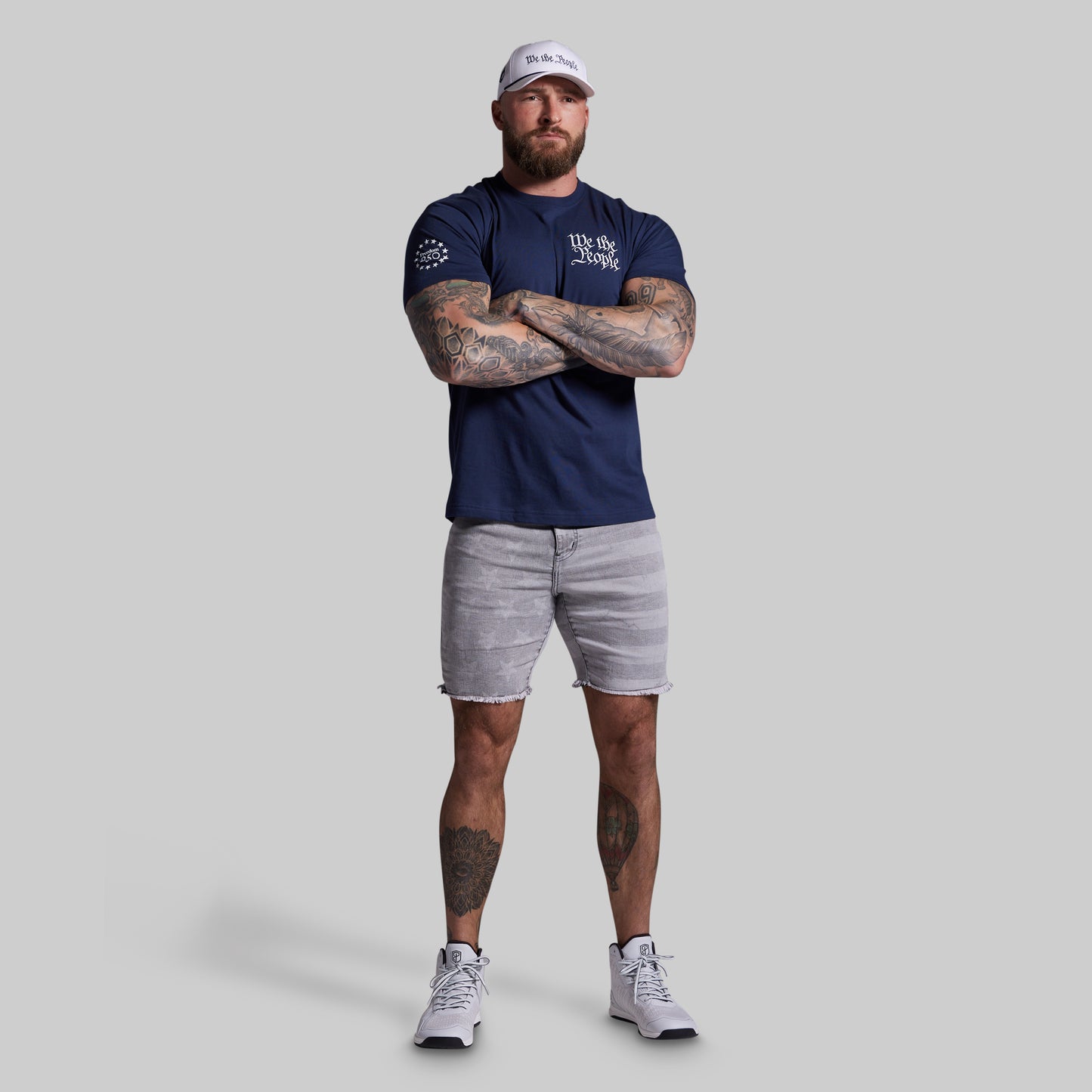 Man wearing a navy blue t-shirt and light gray shorts with tattoos on arms and legs, standing against a plain background.