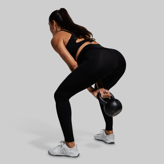 Woman in black sports bra and leggings performing a kettlebell swing against a gray background