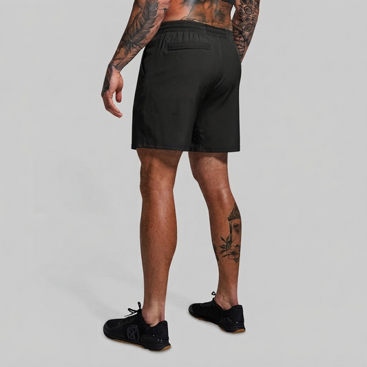 Rear view of a man wearing green athletic shorts and black sneakers against a light gray studio background.