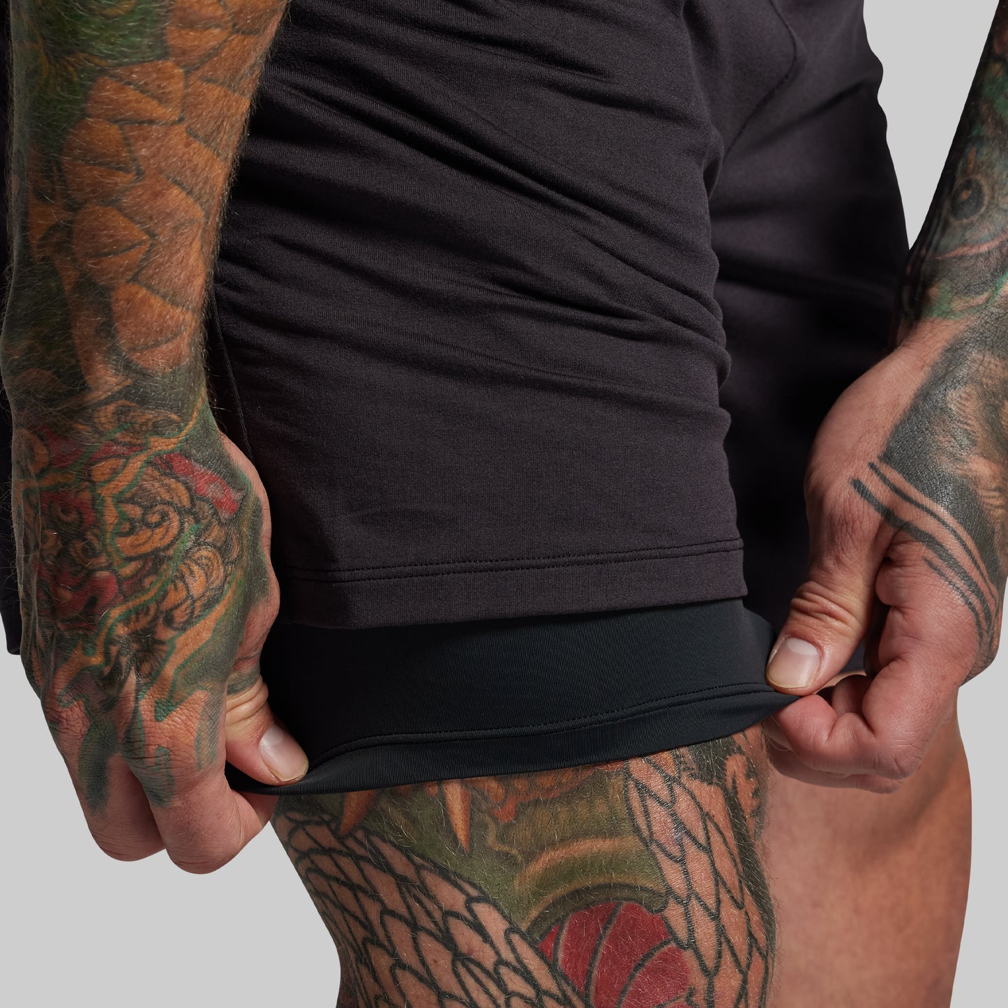 Person wearing a black shorts adjusting compression liner with tattooed arms on a plain background