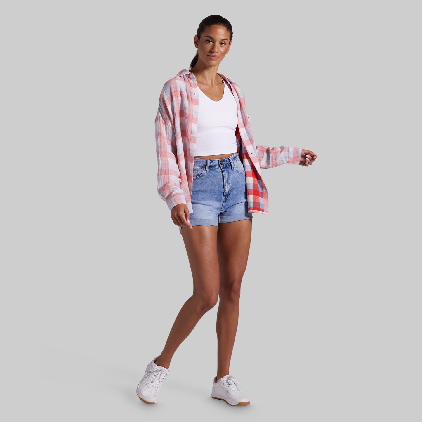 A woman modeling a pink and white plaid button-up shirt over a white crop top and light blue denim shorts, paired with white sneakers, against a solid gray background.
