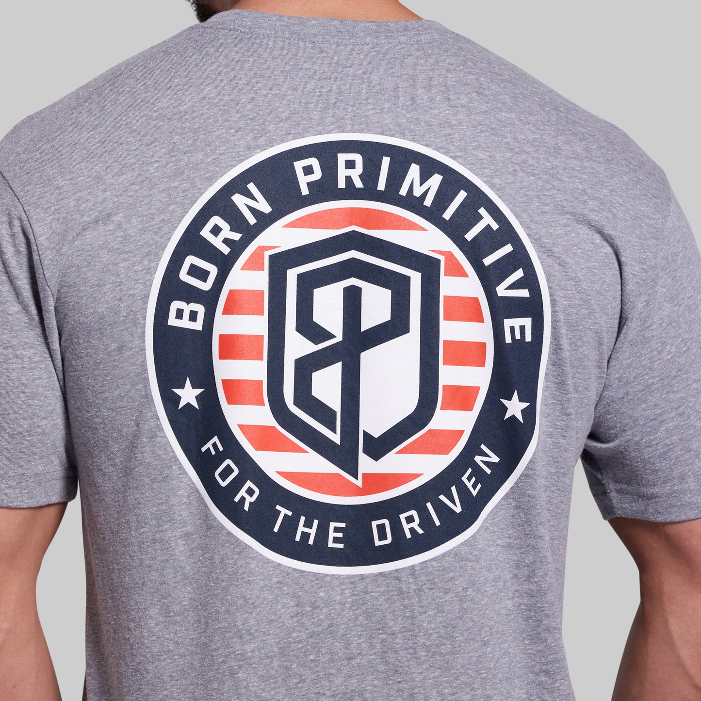 A close-up of a heather gray t-shirt showing the back with a large circular graphic. The graphic features the text 'BORN PRIMITIVE FOR THE DRIVEN' in white around a shield logo with red and white stripes and a navy blue 'BP' monogram in the center.