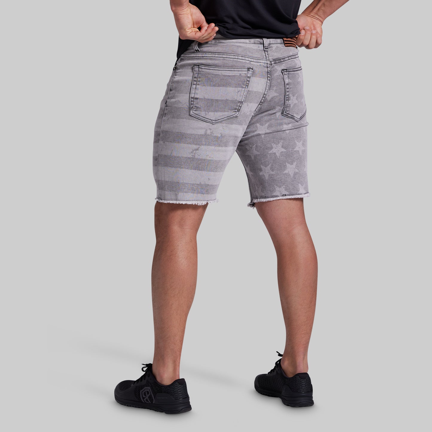 A person shown from the waist down wearing faded gray denim shorts with a distressed American flag pattern, paired with a black shirt and black sneakers, against a solid gray studio background.