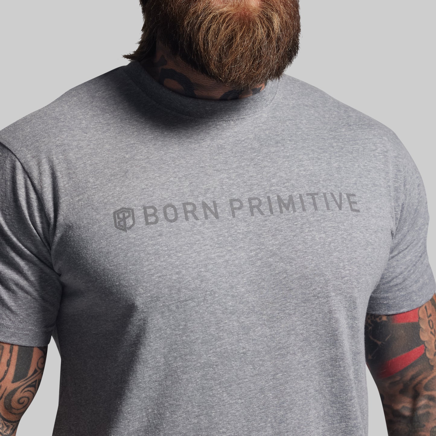 Person wearing a gray 'Born Primitive' t-shirt with tattoos visible on arms.