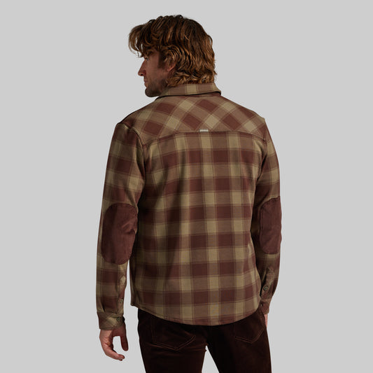 A man shown from the back wearing a brown and beige plaid flannel shirt with brown corduroy elbow patches, set against a solid gray background.