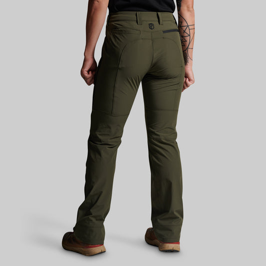 A person shown from the back wearing olive green technical pants, a black shirt, and brown shoes against a light gray studio background.