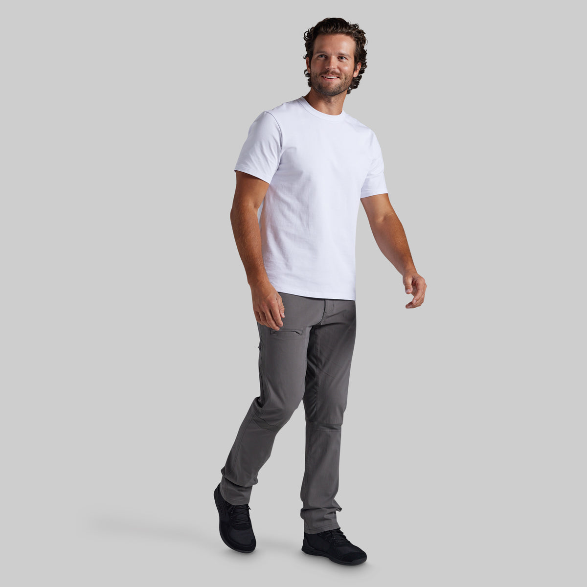 A man modeling a white short-sleeve crewneck t-shirt and gray pants against a solid gray background.
