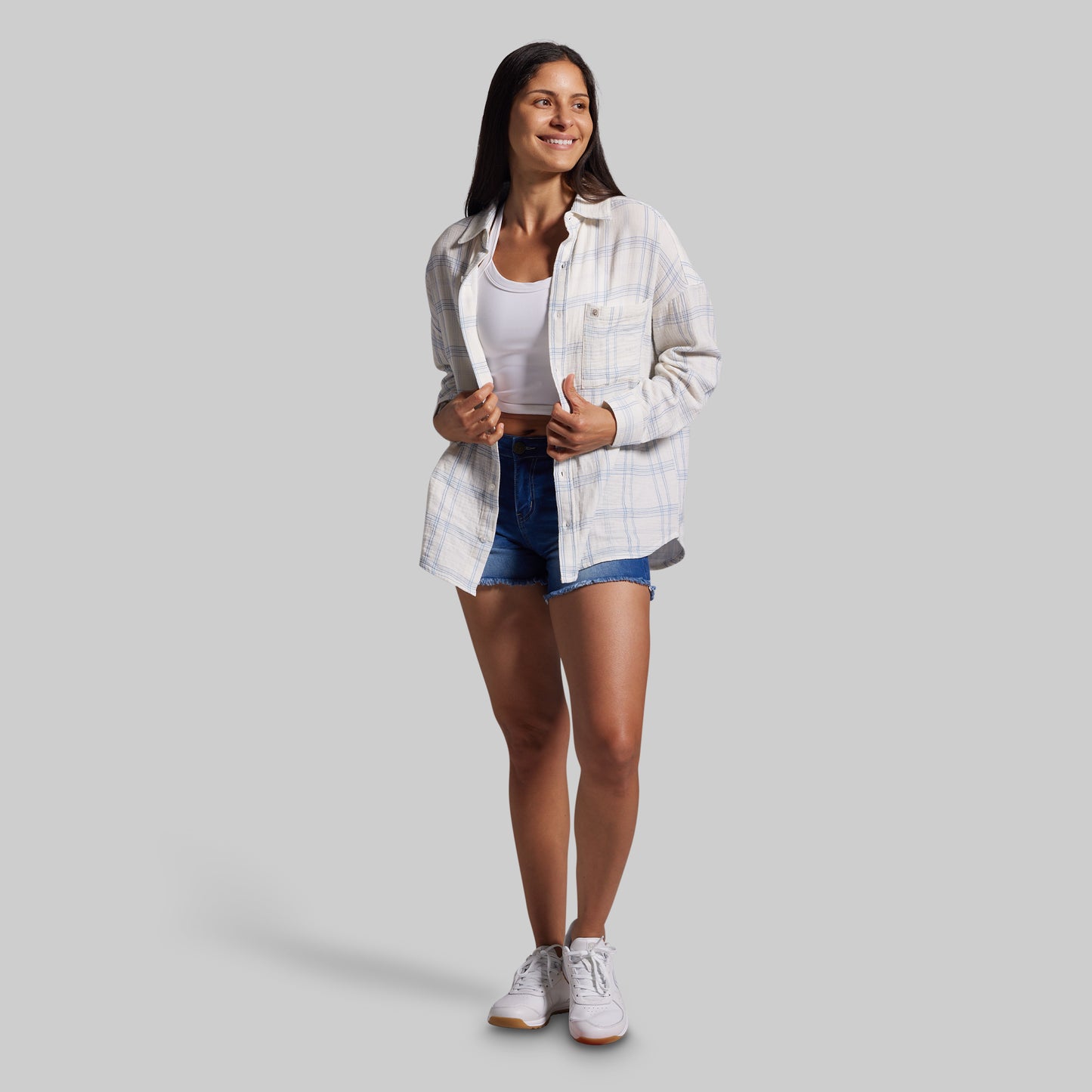Woman wearing a white plaid shirt, white tank top, blue shorts, and white sneakers on a gray background