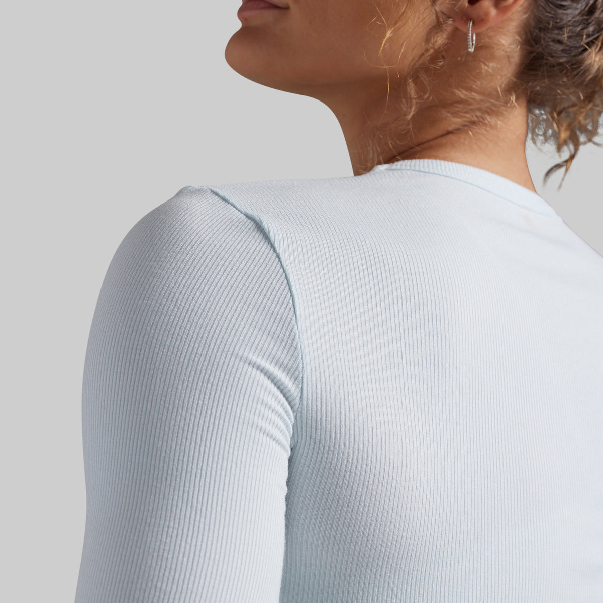 Close-up of a woman wearing a light icy blue ribbed long-sleeve top, showing the back and shoulder detail against a neutral gray background.