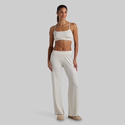 A woman modeling a matching two-piece lounge set consisting of a white lounge bra and white wide-leg pants, standing against a solid light gray background.