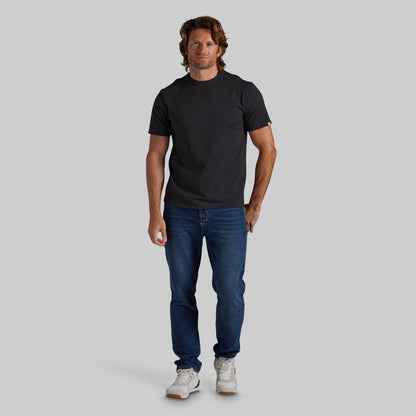 A man modeling a plain black short-sleeve crewneck t-shirt, paired with blue jeans and white sneakers, standing against a solid light gray background.