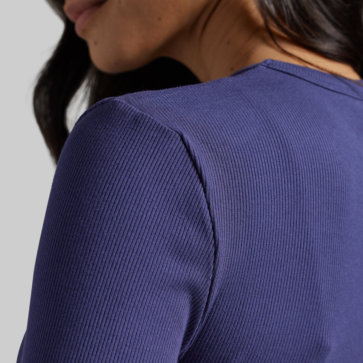 Close-up of a woman wearing a ribbed long-sleeve top in a deep periwinkle or indigo purple color, showing the texture of the fabric and the shoulder seam.