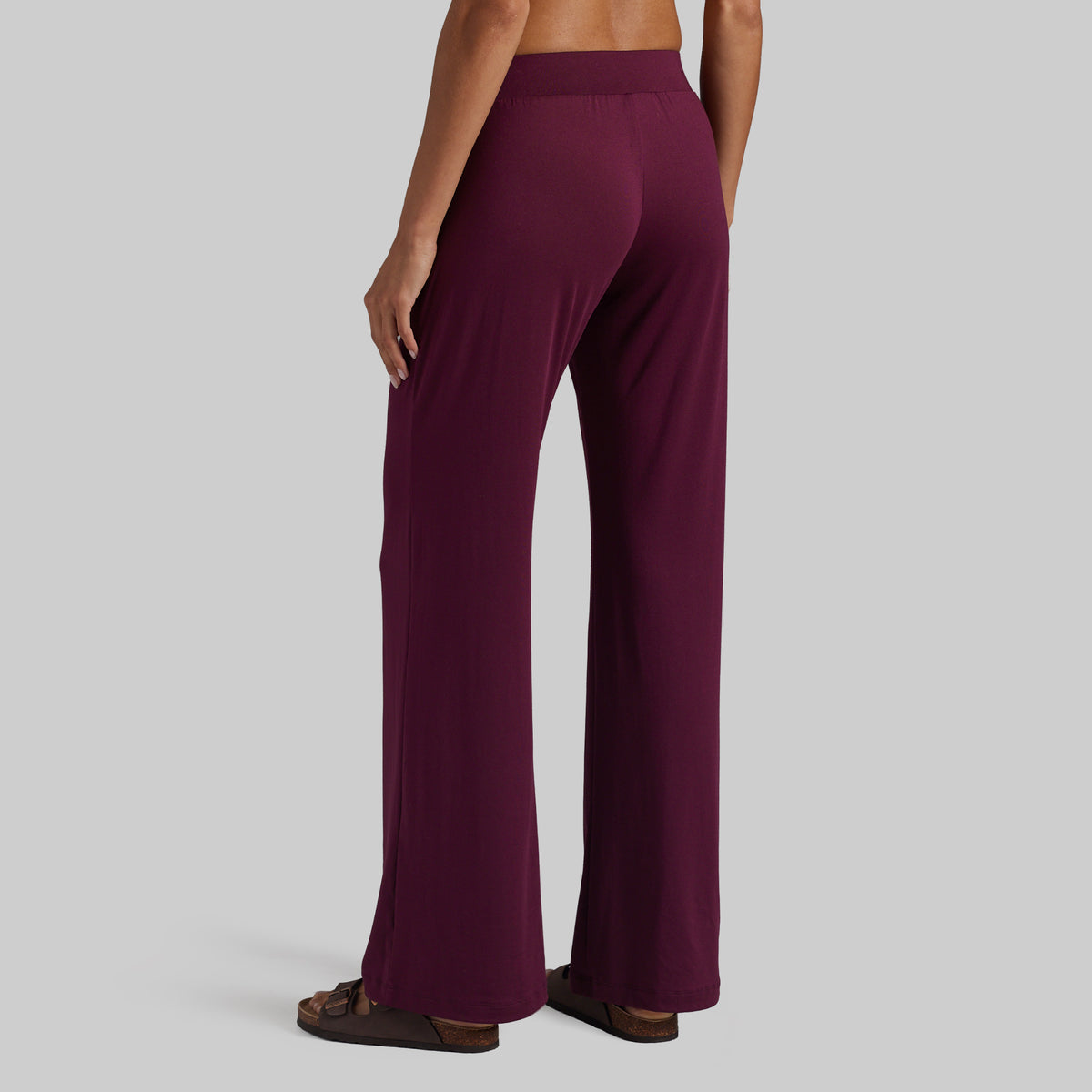 A person shown from the back wearing loose, wide-leg pants in a deep burgundy or plum color, paired with brown sandals, against a plain light gray studio background.