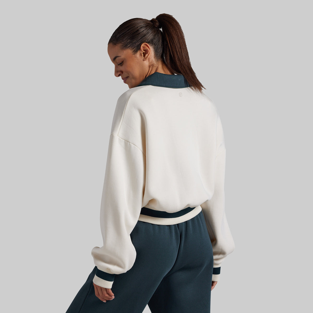 A woman shown from the back wearing a cream-colored cropped sweatshirt with dark teal ribbed trim and matching dark teal sweatpants, set against a solid light gray background.