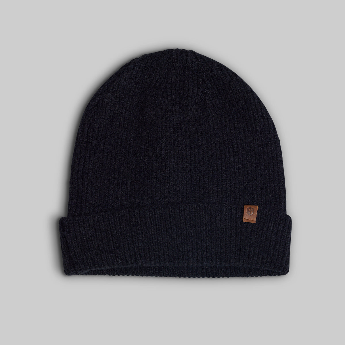 A dark navy blue ribbed knit beanie hat with a small brown leather brand patch on the cuff, shown against a solid gray background.