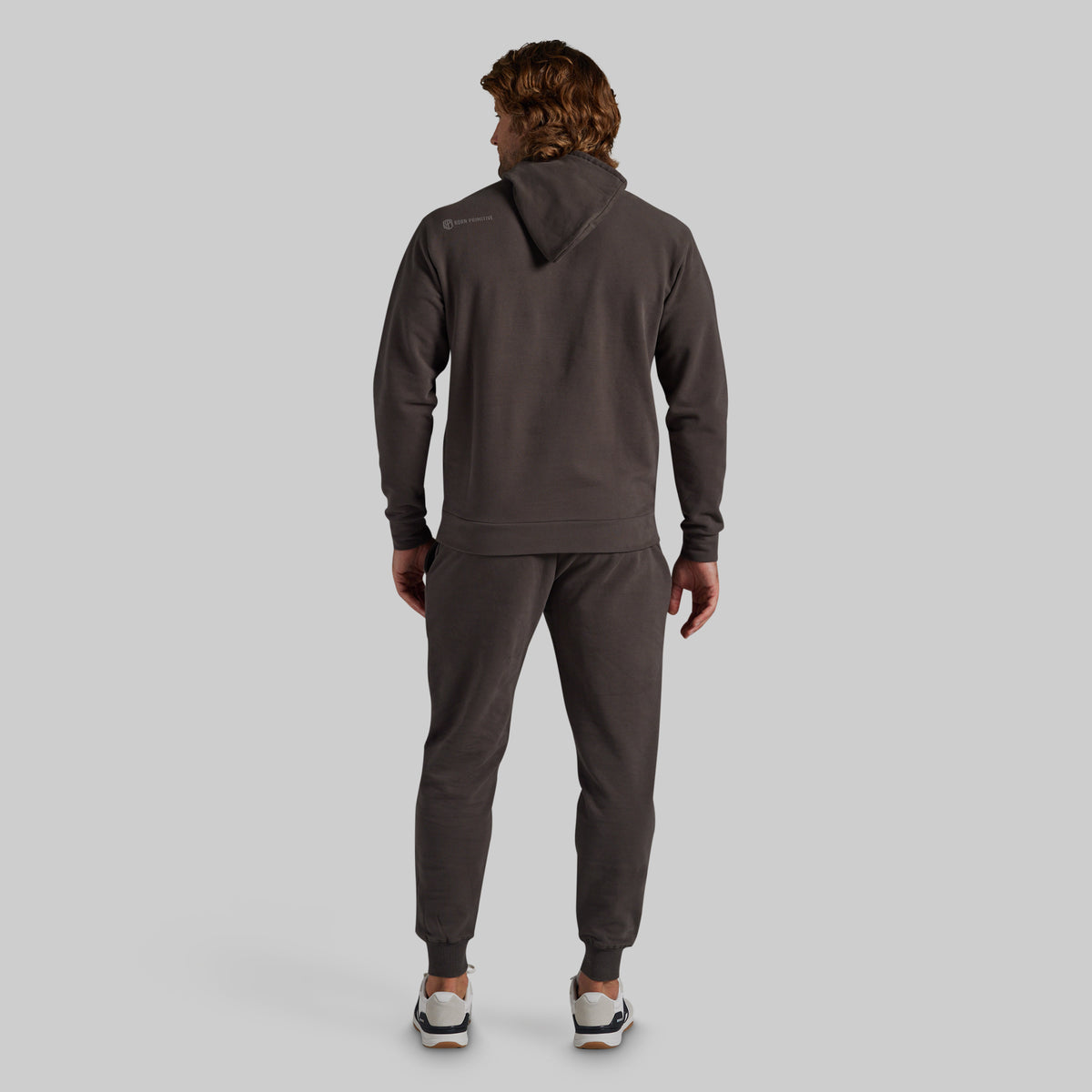 A man viewed from behind wearing a dark grey athletic tracksuit, consisting of a pullover hoodie and matching jogger pants, against a solid light gray background.