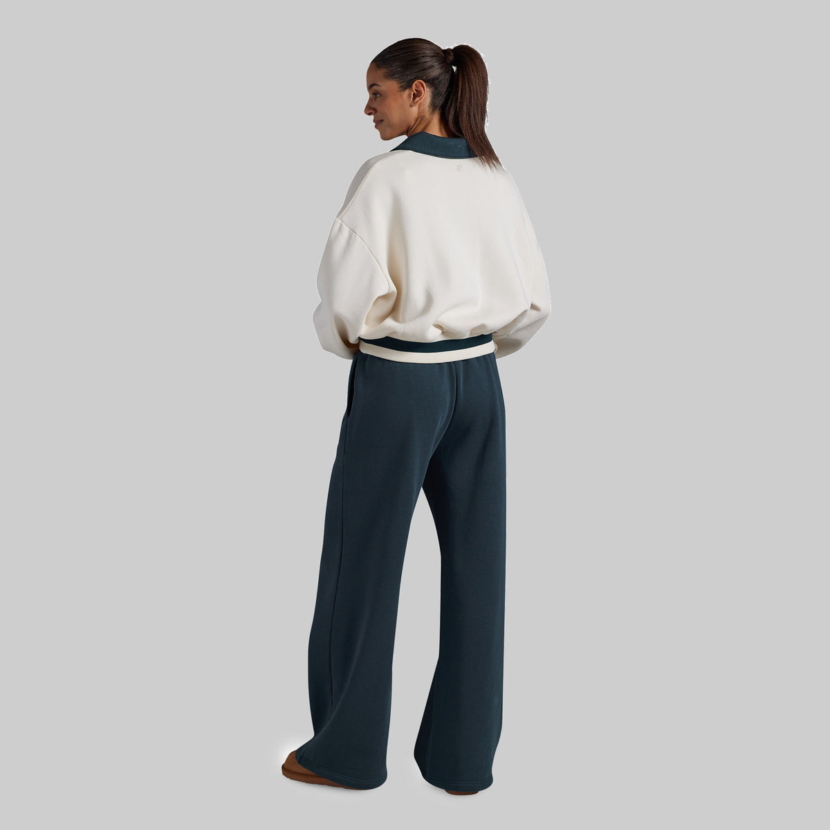 A woman shown from the back wearing a cream sweatshirt with dark teal ribbed trim and matching dark teal wide-leg pants, photographed against a plain light gray studio background.