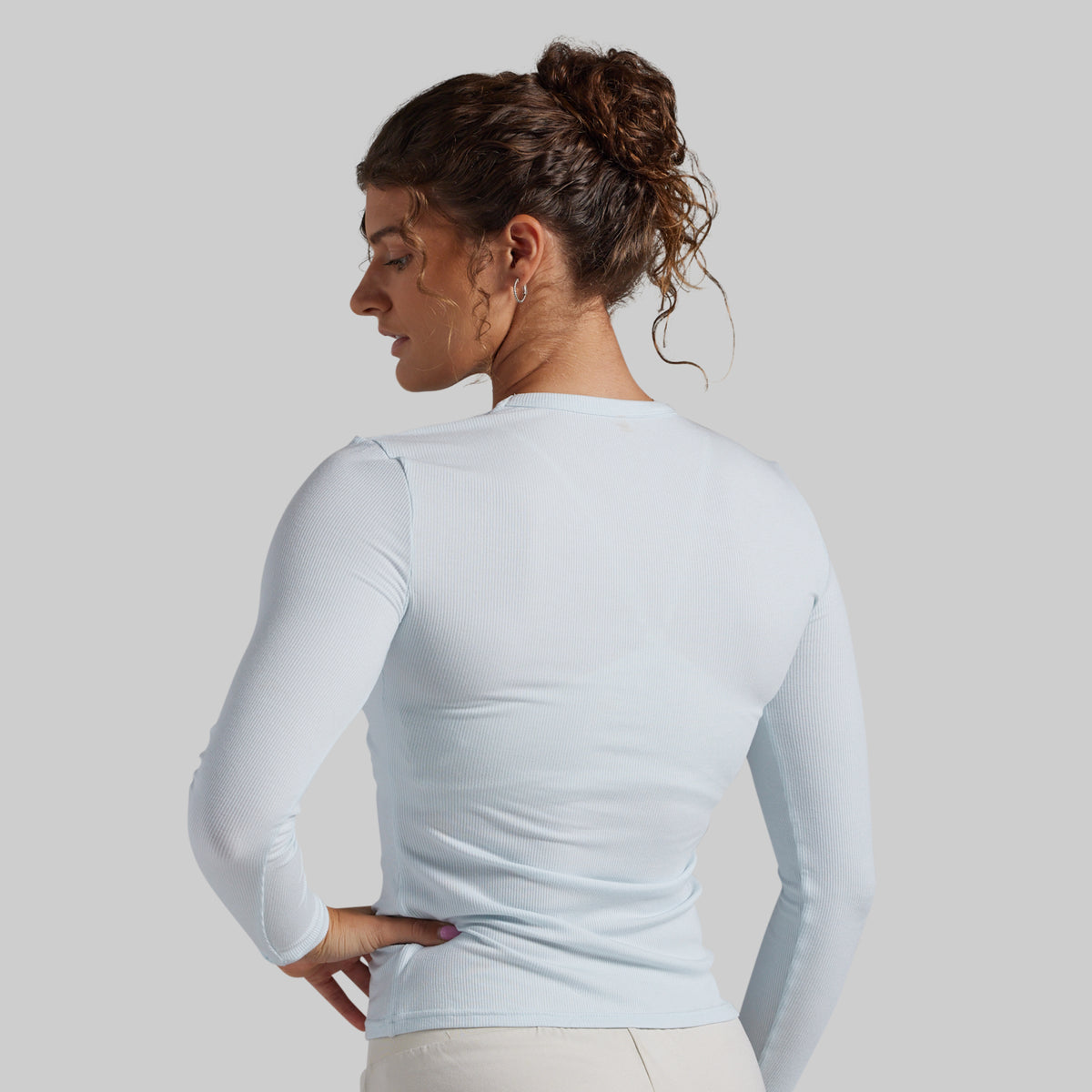 A woman shown from the back wearing a fitted long-sleeve top in a very light icy blue color, paired with light beige pants, against a solid light gray studio background.