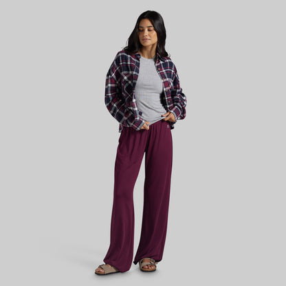 A woman modeling a pair of wide-leg burgundy pants, styled with a gray t-shirt, a dark red and navy plaid flannel shirt, and brown sandals, against a solid gray studio background.