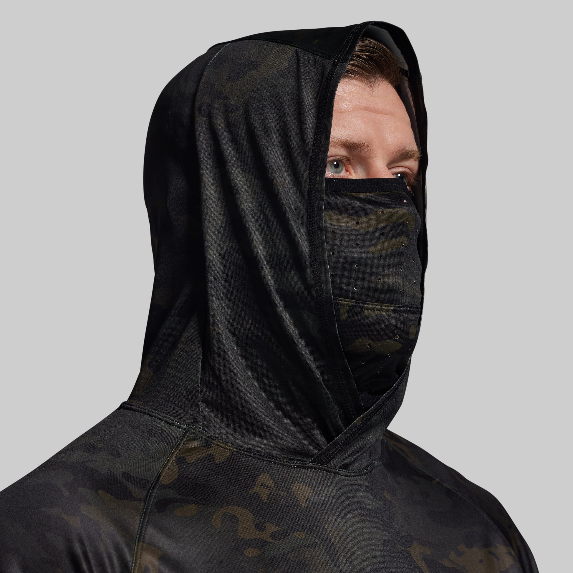 Person wearing a black hooded sun shirt with camouflage pattern and face mask on a gray background