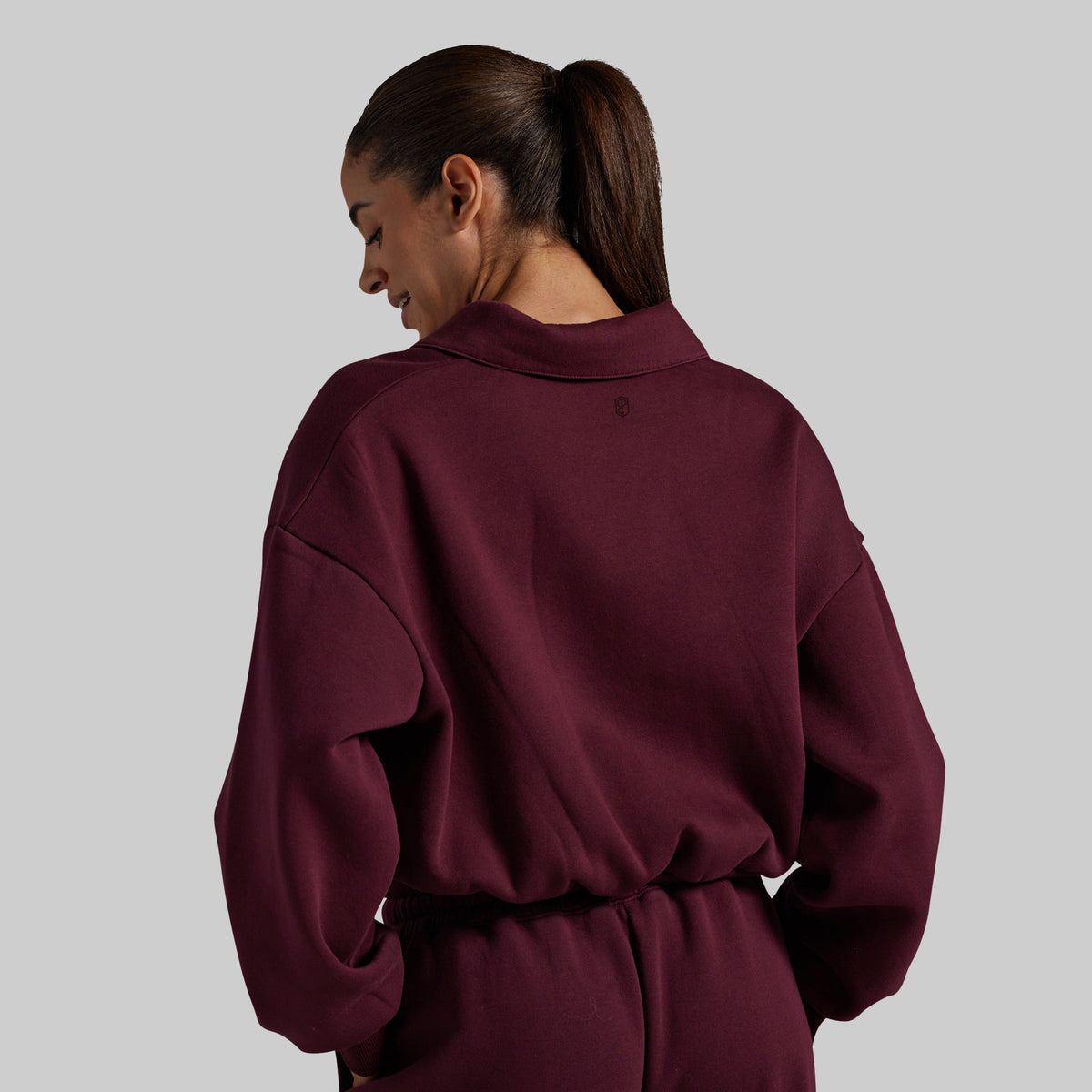 A woman shown from the back wearing a deep burgundy crewneck sweatshirt and matching drawstring sweatpants against a solid gray studio background.