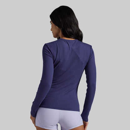 A woman shown from the back wearing a fitted long-sleeve athletic top in a deep purple shade, paired with light lavender shorts, against a solid light gray background.